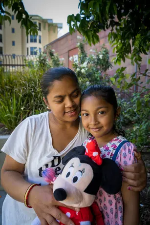 A woman holds a young girl close under a leafy tree, the girl clutching a Minnie Mouse doll and looking straight at the camera while the woman looks down at her, smiling.