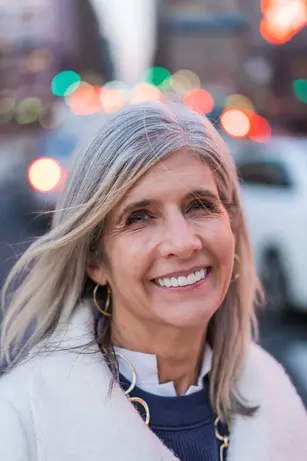 A woman with long grey-blonde hair smiles broadly on a city street at dusk, out-of-focus traffic lights blooming red and green behind her.