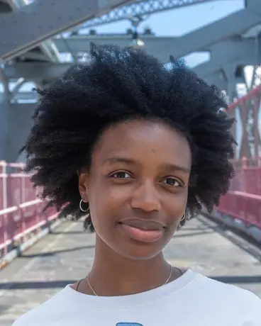 A woman with a natural afro stands on the Williamsburg Bridge walkway, its red railings receding behind her.