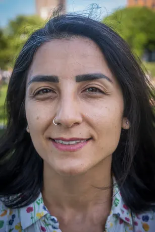 A woman with dark hair and a nose ring smiles gently in a park, greenery and a brick building blurred behind her.