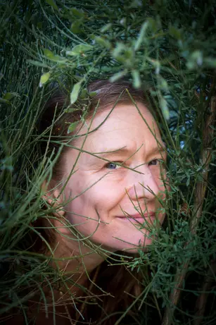 A woman's face framed by a dense tangle of green branches, one eye catching the light as she smiles through the foliage.