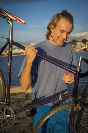 A man with a top knot and beaded earrings laughs as he lifts a purple road bike by its frame, a harbour and moored boats behind him in low golden light.