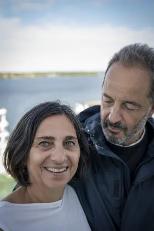 A smiling woman with short dark hair stands in front of a man in a navy jacket who looks down at her, open water and a distant shoreline behind them.