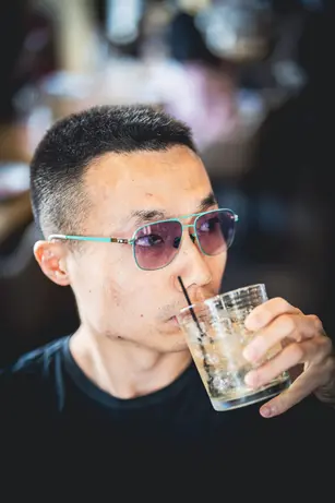 A man in teal-rimmed sunglasses drinks through a straw from a tumbler of ice, the bar behind him falling away into warm blur.
