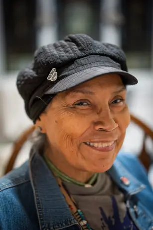 An older woman in a quilted cap and denim jacket turns toward the camera with a warm, crinkled smile.