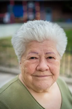 An older woman with a shock of white hair and a green top gives a small, closed-mouth smile on a residential street.