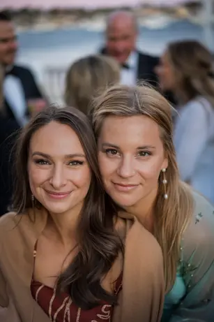 Two women lean cheek to cheek at an outdoor evening event, formally dressed, other guests blurred behind them.