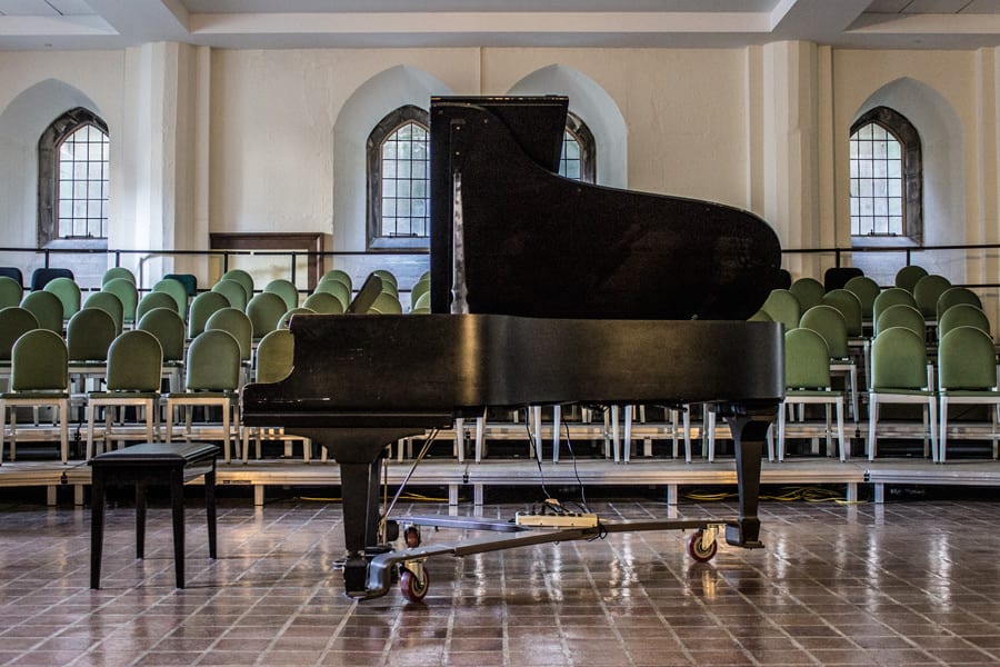 A black grand piano on castors at the front of the chapel lecture room, rows of green-upholstered chairs behind it and arched leaded windows along the wall.