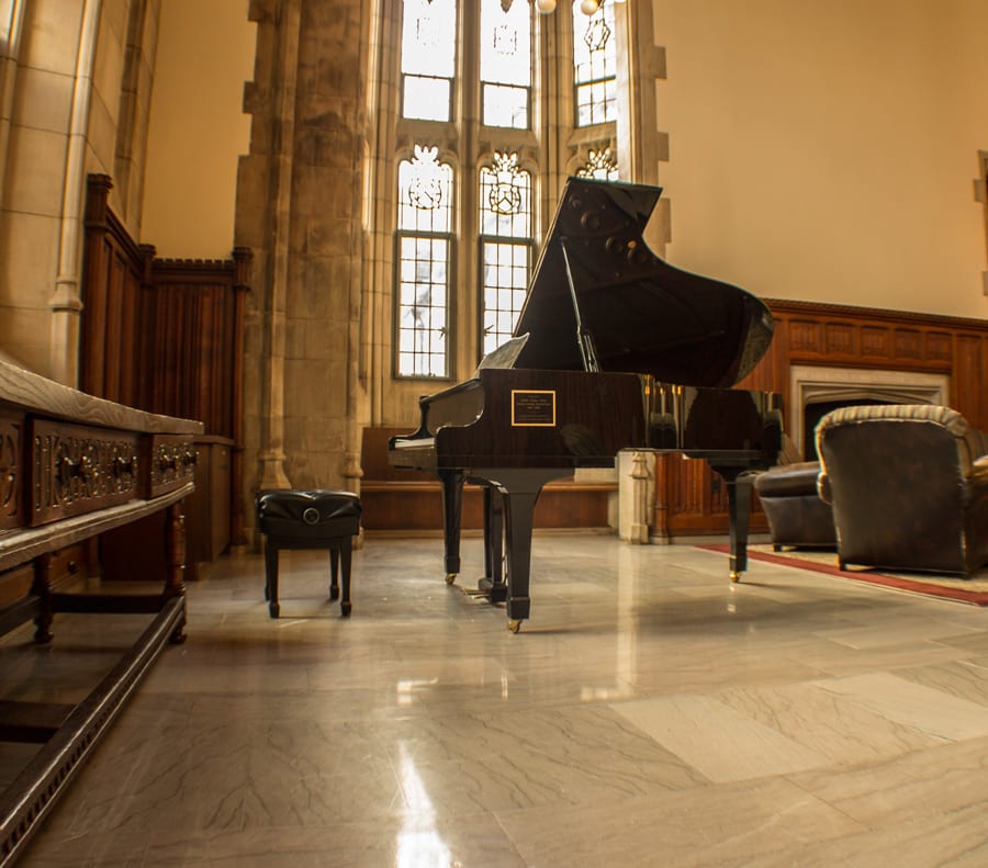 A black grand piano in a wood-panelled common room, lit by a tall gothic window, with leather armchairs beside it.
