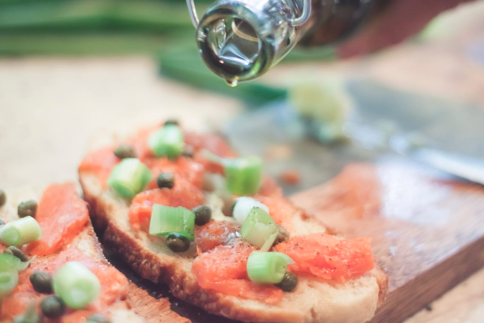 Olive oil poured from a bottle over two assembled open-faced sandwiches, salmon, capers and diced green onion already on the toast.