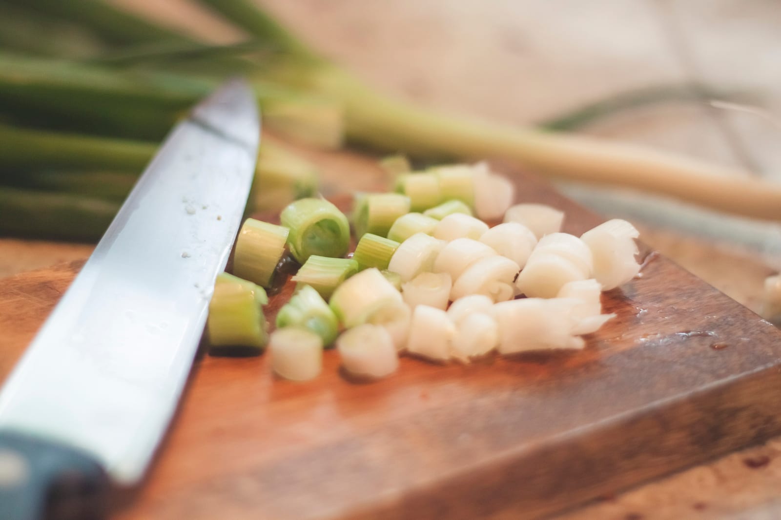 A chef's knife lying on the board next to finely diced green onion, whole onions still bundled behind it.