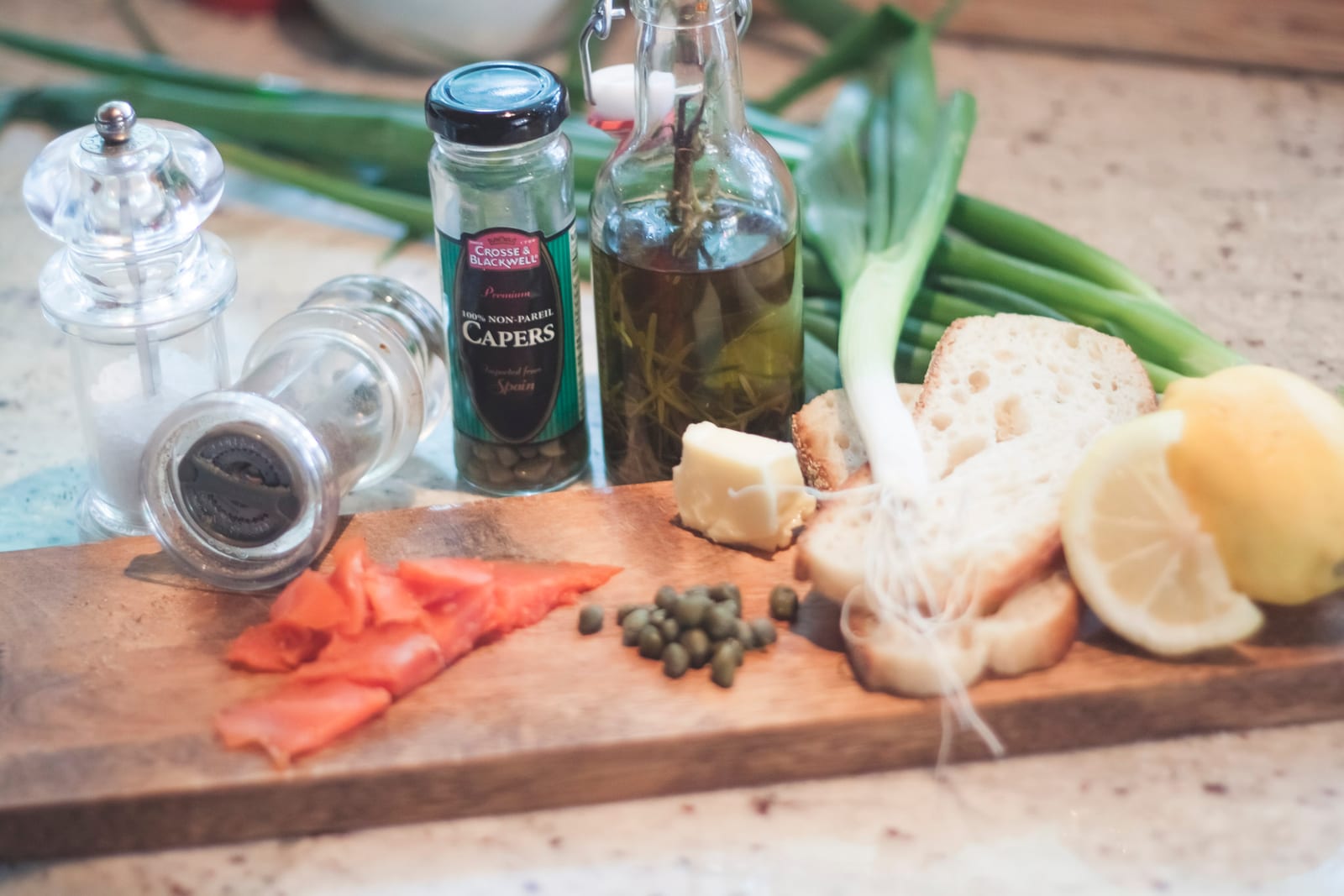 The ingredients laid out on a wooden board: salt and pepper grinders, a jar of capers, a bottle of olive oil, a bundle of green onions, butter, sliced bread, lemon halves and a few slices of smoked salmon.
