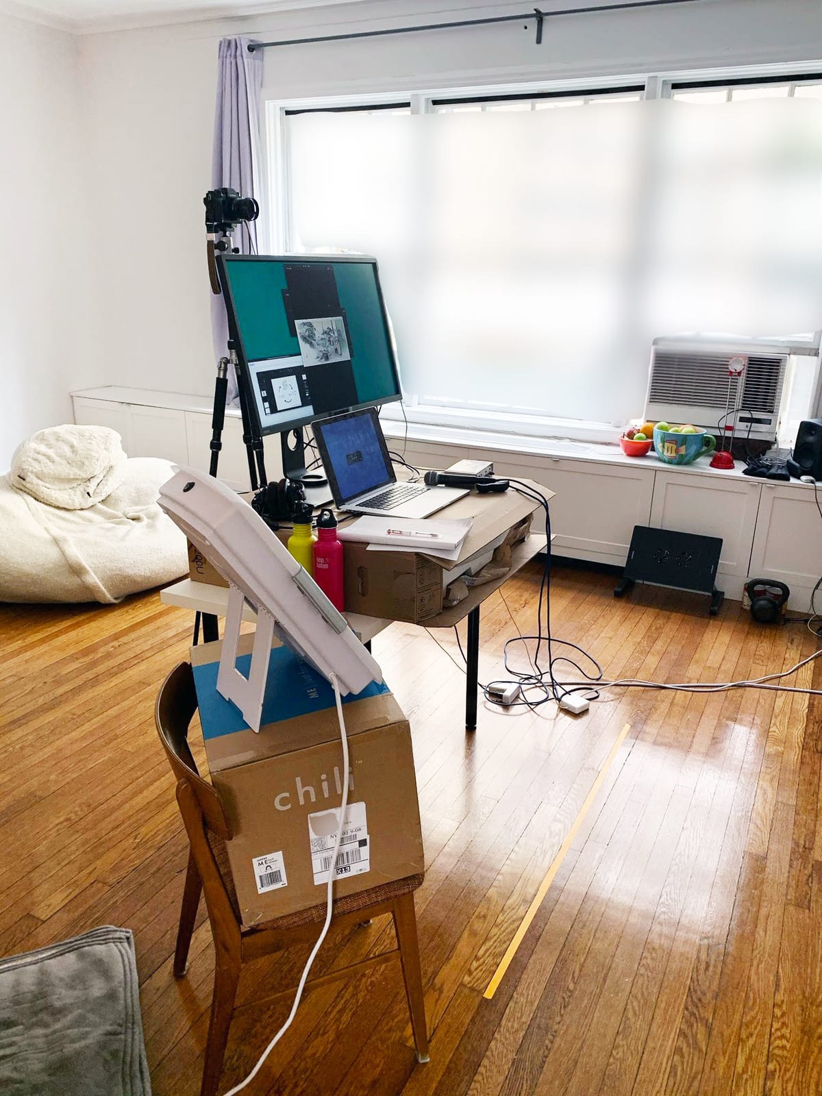 A makeshift home recording rig: an external monitor showing a slide, a camera on a tripod just above it, a laptop on a desk beside a stack of boxes, a microphone on a boom arm in the foreground, and cables running across a wood floor.